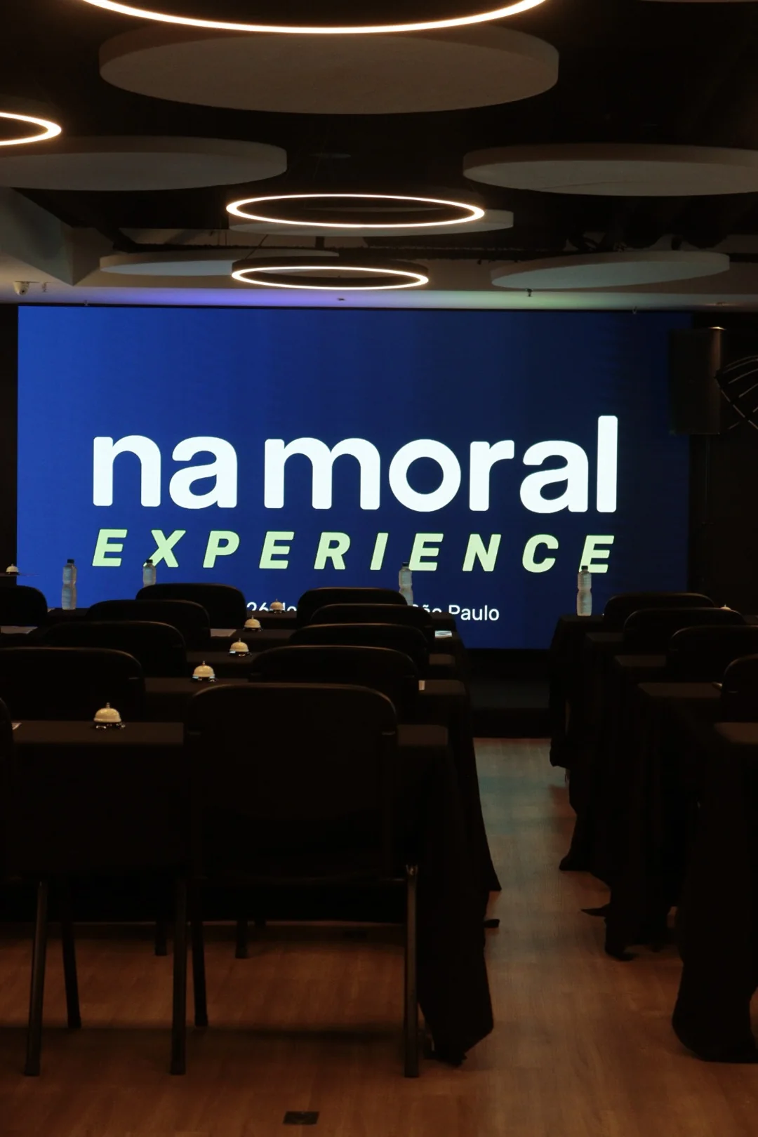Ambiente do Na Moral Experience, com mesas e a marca do evento no palco